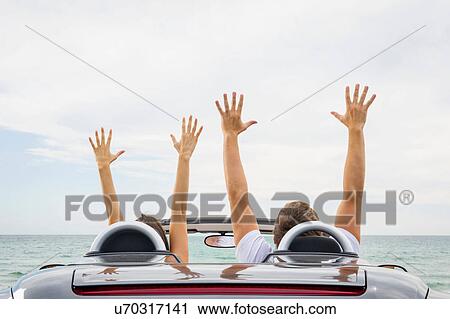 Stock Image - Rear view of young couple sitting in convertible sports car, arms raised. Fotosearch