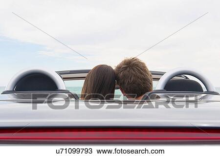 Stock Image - Rear view of young couple sitting in convertible sports car. Fotosearch