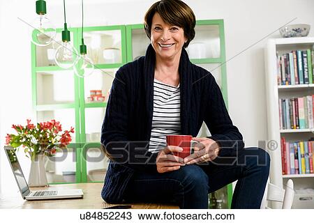 Stock Image - Senior woman sitting on desk holding mug. Fotosearch