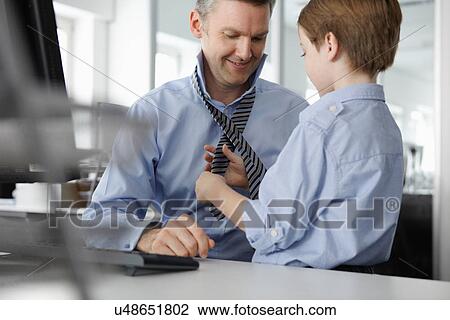 Stock Image - Son putting tie on father at desk. Fotosearch