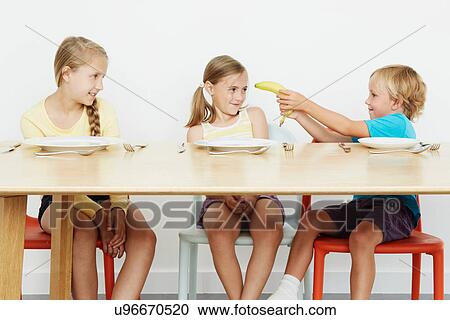 Three children at table, boy holding banana View Large Photo Image Stock Image - Three children at table, boy holding banana. Fotosearch