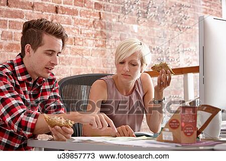Stock Image - Two colleagues eating sandwiches at desk. Fotosearch