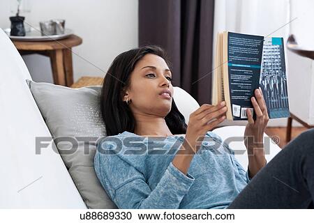 Stock Image - Woman reclining on sofa reading book. Fotosearch