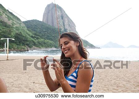 Stock Image - Woman using camera phone on beach, Rio de Janeiro, Brazil. Fotosearch