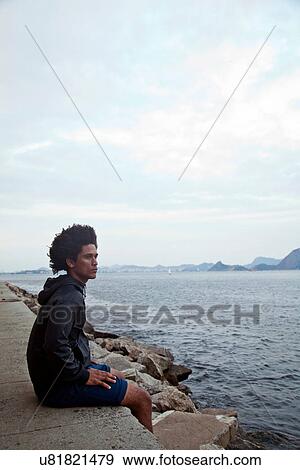 Young man sitting on wall looking at sea, Rio de Janeiro, Brazil View Large Photo Image Stock Photo - Young man sitting on wall looking at sea, Rio de Janeiro, Brazil. Fotosearch
