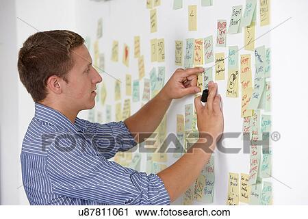 Stock Image - Young man sticking adhesive note on wall. Fotosearch