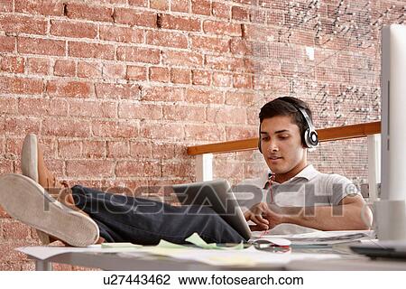 Stock Image - Young man using digital tablet wearing headphones, feet up. Fotosearch