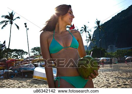 Stock Image - Young woman holding coconut drink on beach. Fotosearch