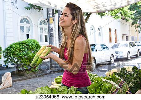 Stock Photography - Young woman holding corn on the cob in market. Fotosearch
