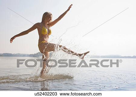 Stock Image - Young woman kicking water in lake. Fotosearch
