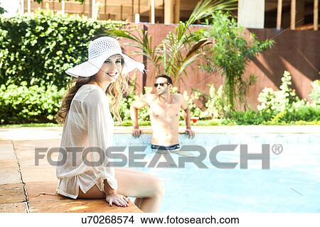 Picture - Young woman wearing sunhat sitting on poolside. Fotosearch