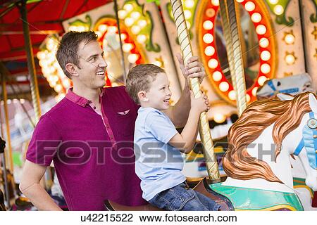 Stock Image - Father and son (4-5) on carousel in amusement park. Fotosearch