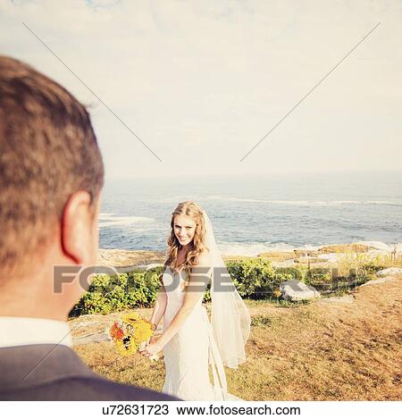 Stock Image - Groom looking at bride, sea in background. Fotosearch