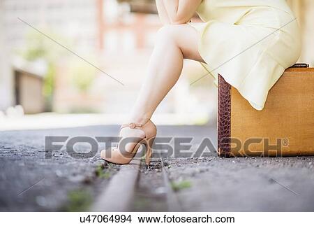 Picture - Low section of woman in dress sitting on suitcase at train station. Fotosearch