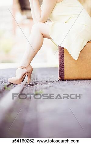 Stock Image - Low section of woman in dress sitting on suitcase at train station. Fotosearch