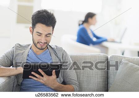 Man sitting on sofa using digital tablet at home, woman in background View Large Photo Image Stock Image - Man sitting on sofa using digital tablet at home, woman in background. Fotosearch