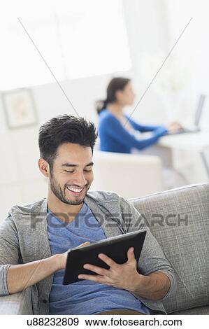 Man sitting on sofa using digital tablet at home, woman in background View Large Photo Image Stock Photo - Man sitting on sofa using digital tablet at home, woman in background. Fotosearch