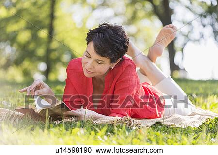Stock Image - Mature woman lying on grass and reading book. Fotosearch