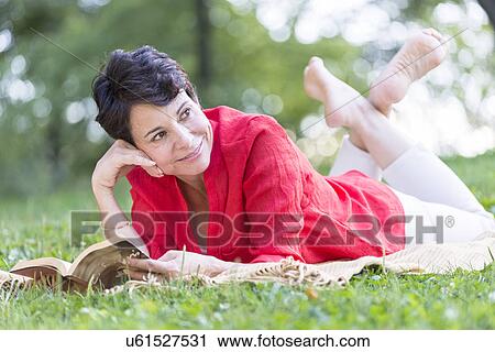 Stock Image - Mature woman lying on grass and reading book. Fotosearch
