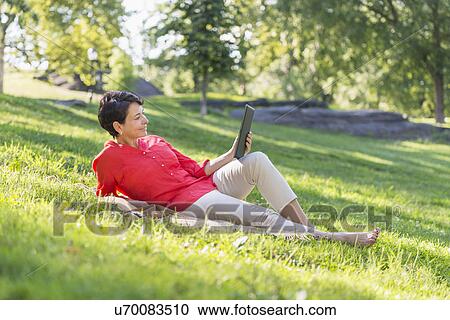 Stock Image - Mature woman lying on grass and using digital tablet. Fotosearch