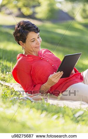 Stock Photo - Mature woman lying on grass and using digital tablet. Fotosearch
