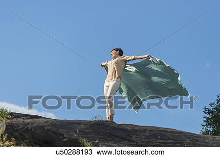 Stock Image - Mature woman standing on rock with arms raised. Fotosearch