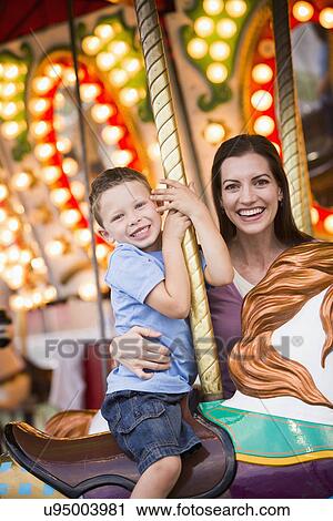 Stock Image - Mother and son (4-5) on carousel in amusement park. Fotosearch