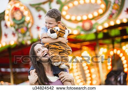 Stock Image - Mother giving her son (4-5) piggyback ride in amusement park. Fotosearch