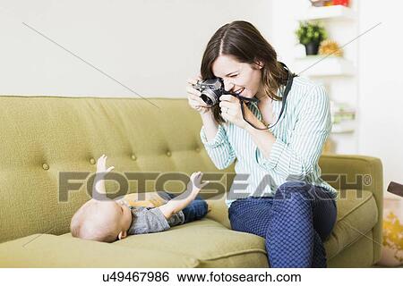 Stock Photograph - Mother sitting on sofa photographing baby boy (6-11 months). Fotosearch