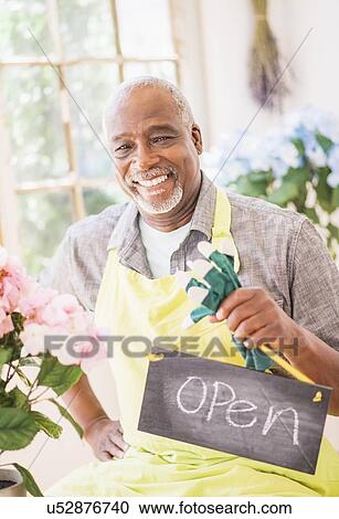 Stock Image - Portrait of man holding open sign in greenhouse. Fotosearch