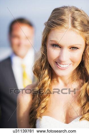 Stock Image - Portrait of smiling bride, groom in background. Fotosearch