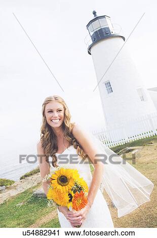 Stock Image - Portrait of smiling bride holding sunflower bouquet, lighthouse in background. Fotosearch