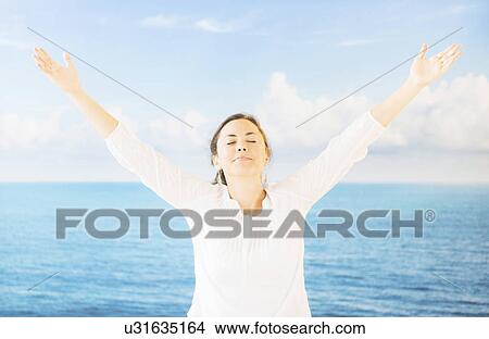 Picture - Portrait of woman with arms raised in front of backdrop with sea and sky. Fotosearch