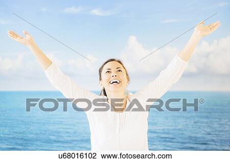Stock Image - Portrait of woman with arms raised in front of backdrop with sea and sky. Fotosearch