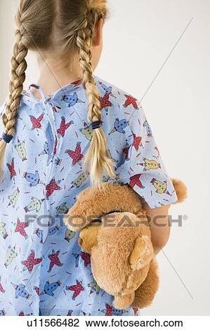 Stock Image - Rear view of blond girl (8-9) in hospital holding teddy bear. Fotosearch