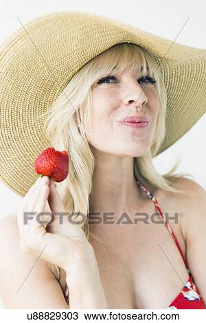 Stock Image - Studio portrait of blonde woman with sun hat holding strawberry. Fotosearch