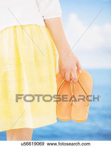 Stock Photo - Studio Shot Mid section of woman holding flip-flops, rear view. Fotosearch