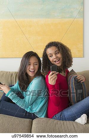 Two friends (12-13, 16-17) sitting on sofa and playing with smartphone View Large Photo Image Picture - Two friends (12-13, 16-17) sitting on sofa and playing with smartphone. Fotosearch