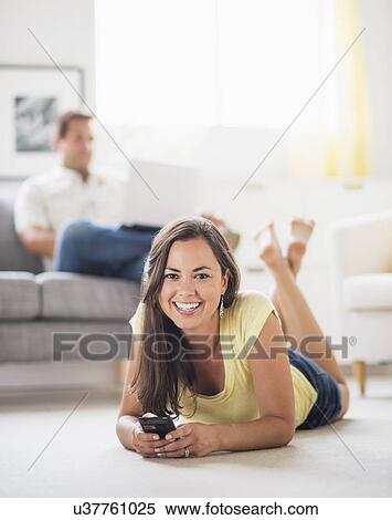 Stock Photography - Woman lying on front and using mobile phone, man in background. Fotosearch