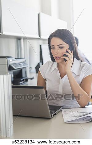 Woman working on laptop and talking on cell phone in office, man in background View Large Photo Image Picture - Woman working on laptop and talking on cell phone in office, man in background. Fotosearch