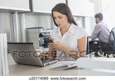 Stock Photo - Woman working on laptop and text messaging in office, man in background. Fotosearch