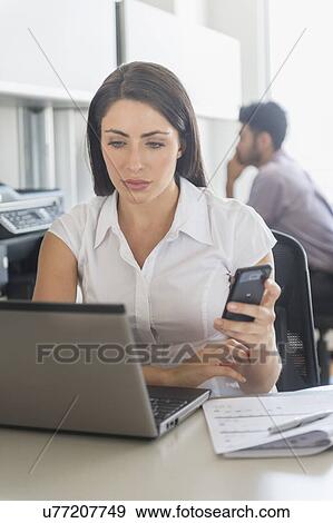 Stock Photo - Woman working on laptop and text messaging in office, man in background. Fotosearch