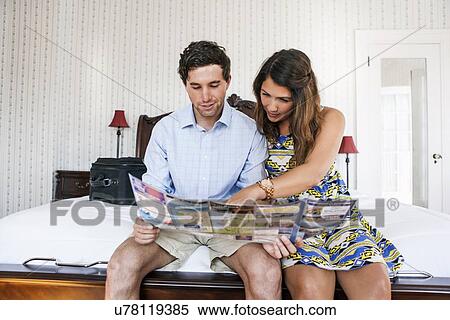 Stock Photography - Young couple sitting on bed in hotel room and reading brochure. Fotosearch