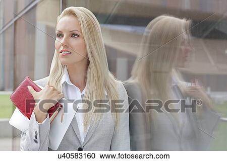 Stock Image - Beautiful businesswoman holding organizer and digital tablet while looking away by glass wall. Fotosearch
