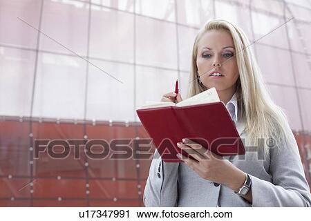 Stock Image - Beautiful businesswoman reading notes in organizer while standing against office building. Fotosearch