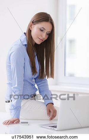 Beautiful businesswoman sitting on desk and looking at laptop View Large Photo Image Stock Image - Beautiful businesswoman sitting on desk and looking at laptop . Fotosearch