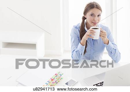 Stock Image - Beautiful woman drinking coffee at her desk. Fotosearch