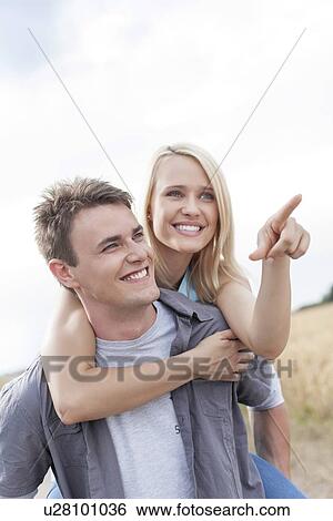 Stock Photograph - Beautiful woman showing something to man while enjoying piggyback ride at field. Fotosearch