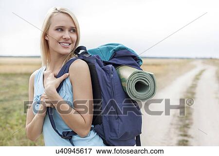 Stock Photo - Beautiful woman with backpack looking away while hiking at field. Fotosearch