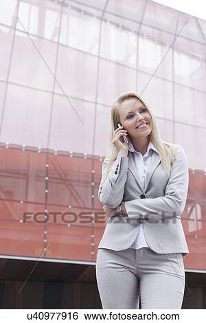 Stock Photograph - Beautiful young businesswoman using mobile phone while standing against office building. Fotosearch